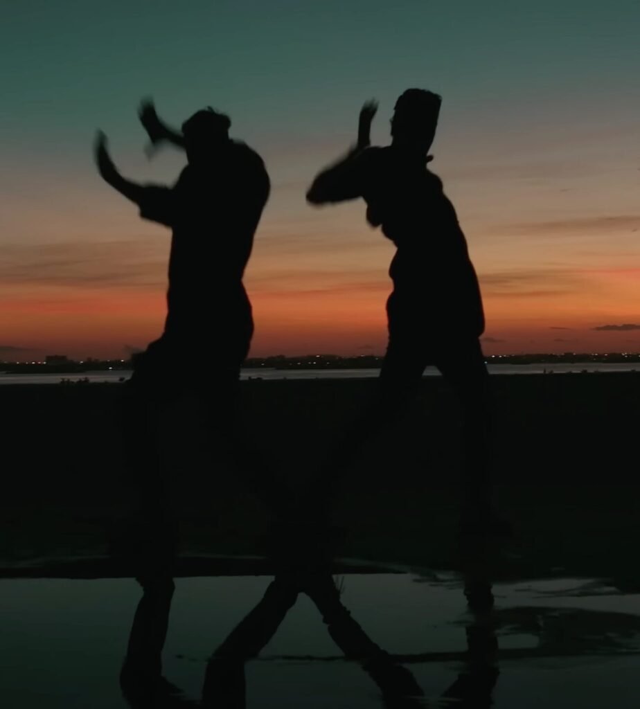 Two people dancing in silhouette on wet ground beneath a fading orange dusk sky